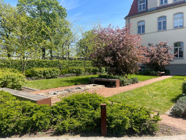 Innenhof mit Grünanlage zum Pausemachen Landschaft mit Bäumen, Blumen und einem gepflasterten Weg vor einem Gebäude.