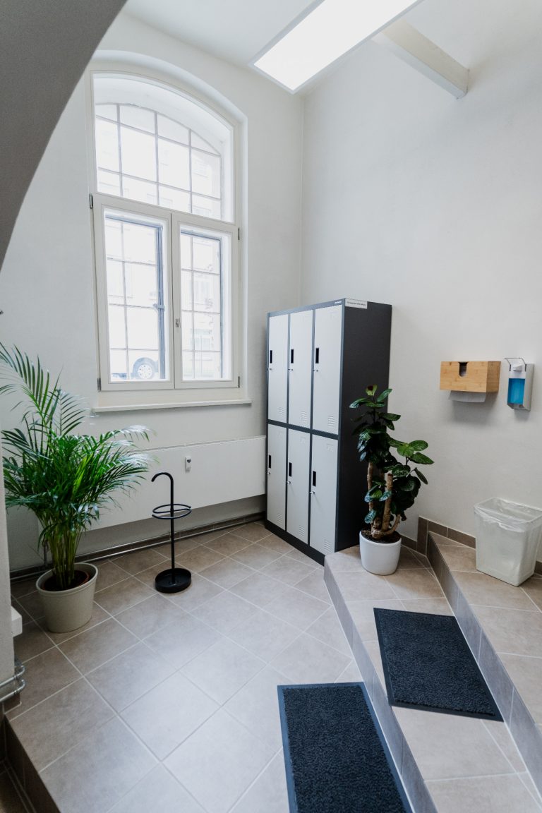 Eingansbereich mit Spind und Händedesinfektion Heller Raum mit Schränken, Pflanzen und einem Treppe zu einer Toilette.