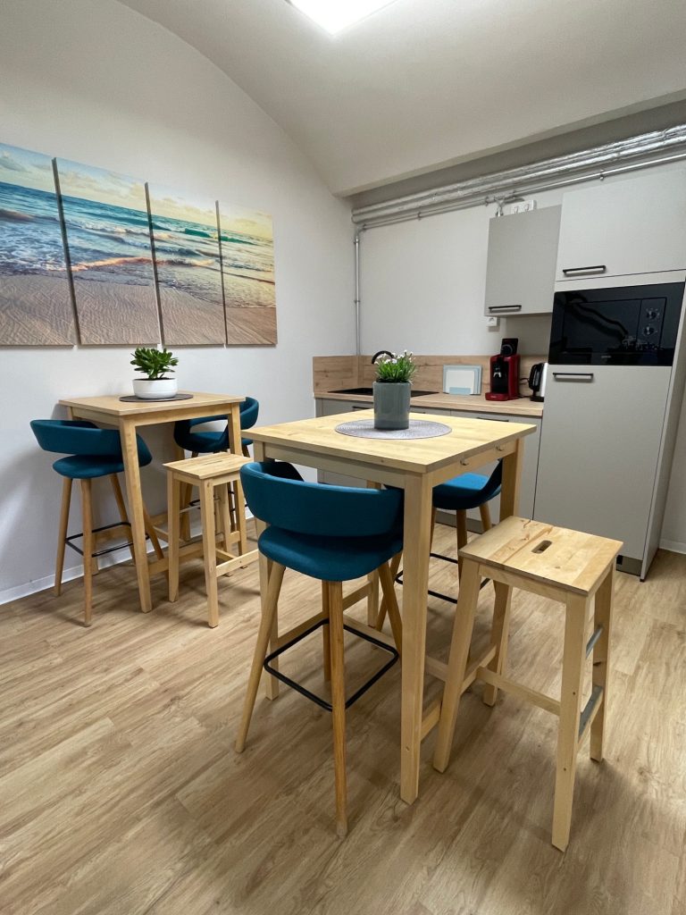 Büroküche mit Kaffeemaschine Moderne Küche mit Holztischen, Stühlen in Blau und Kühlschrank. Wandbild mit Strandmotiv.